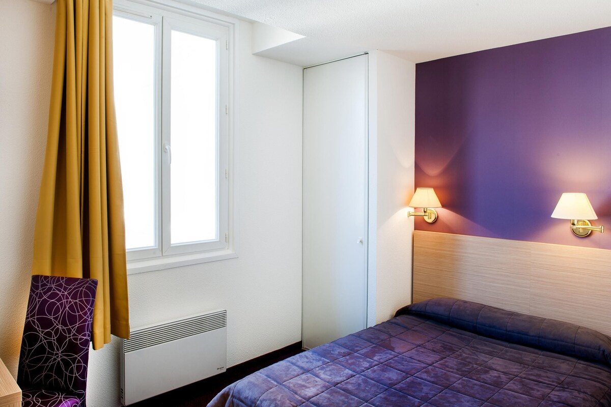 Una habitación de hotel con cama, ventana con cortinas, lámpara de pared y radiador.