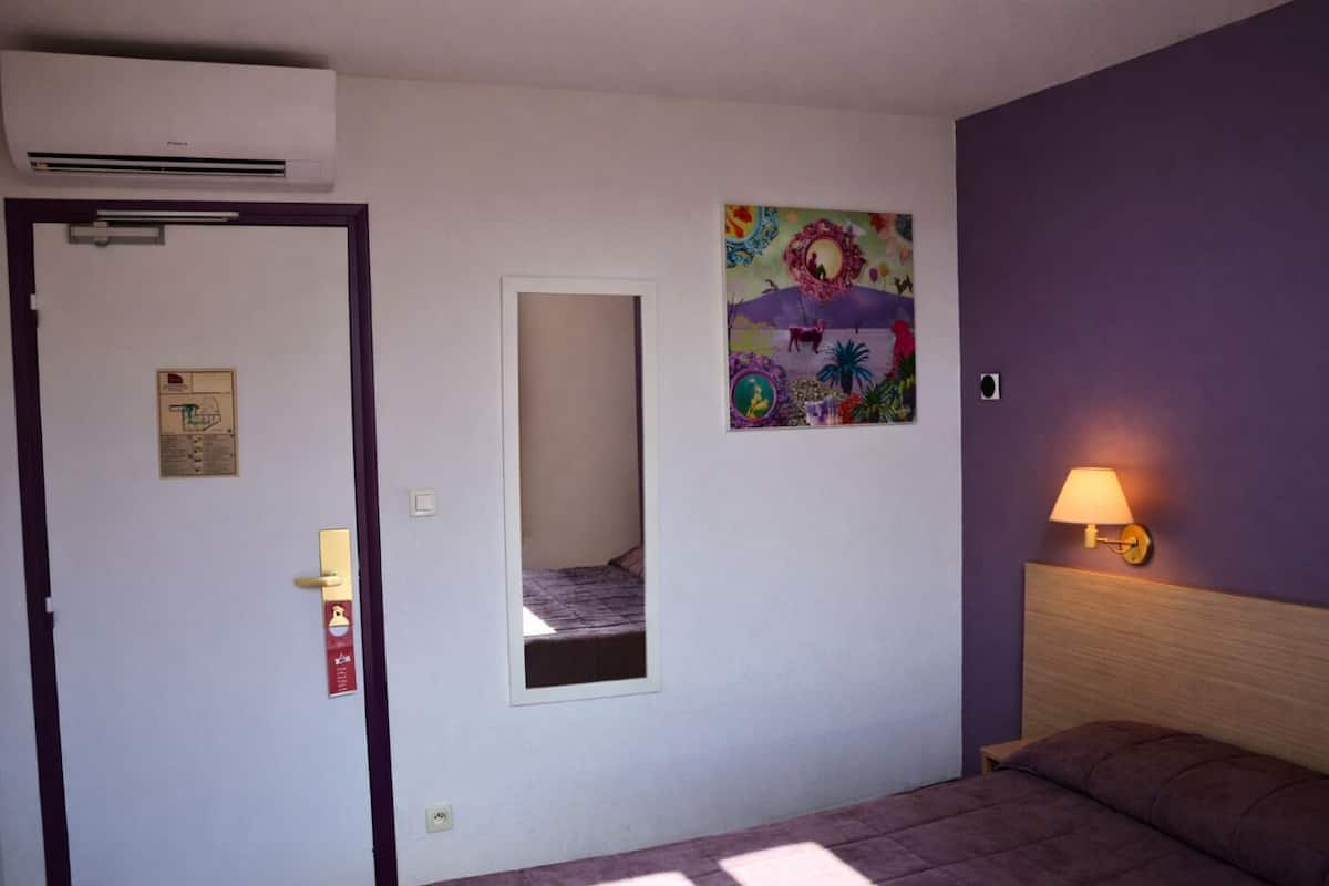 A bedroom with a bed, a wall-mounted lamp, a door with a mirror, and a colorful painting.