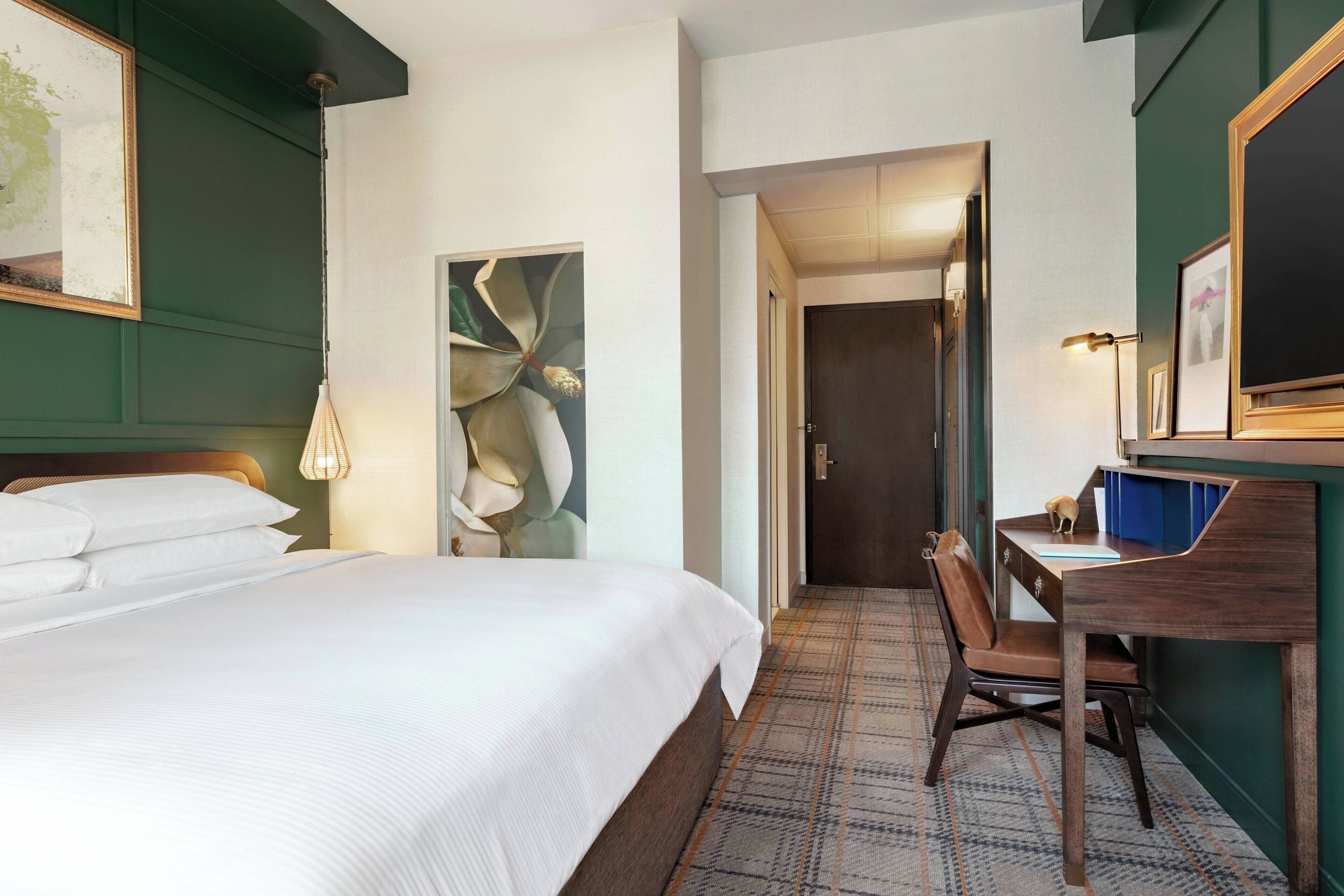 A hotel room with a large bed, a desk, and a chair. There is a door leading to another room.