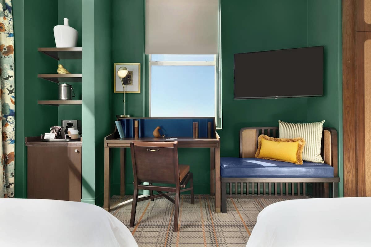 Habitación de hotel con cama, escritorio con silla, televisión, ventana y sofá con cojines.