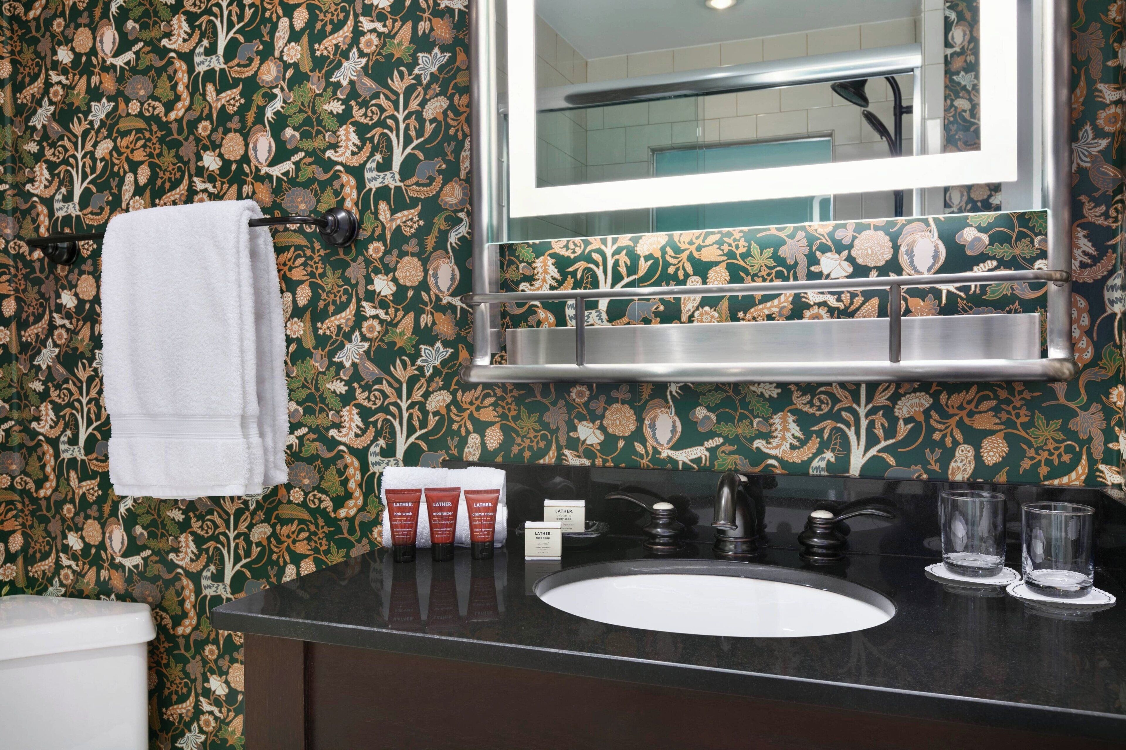 A bathroom with a floral wallpaper, a white towel hanging, a sink with two glasses, and a mirror above the sink.