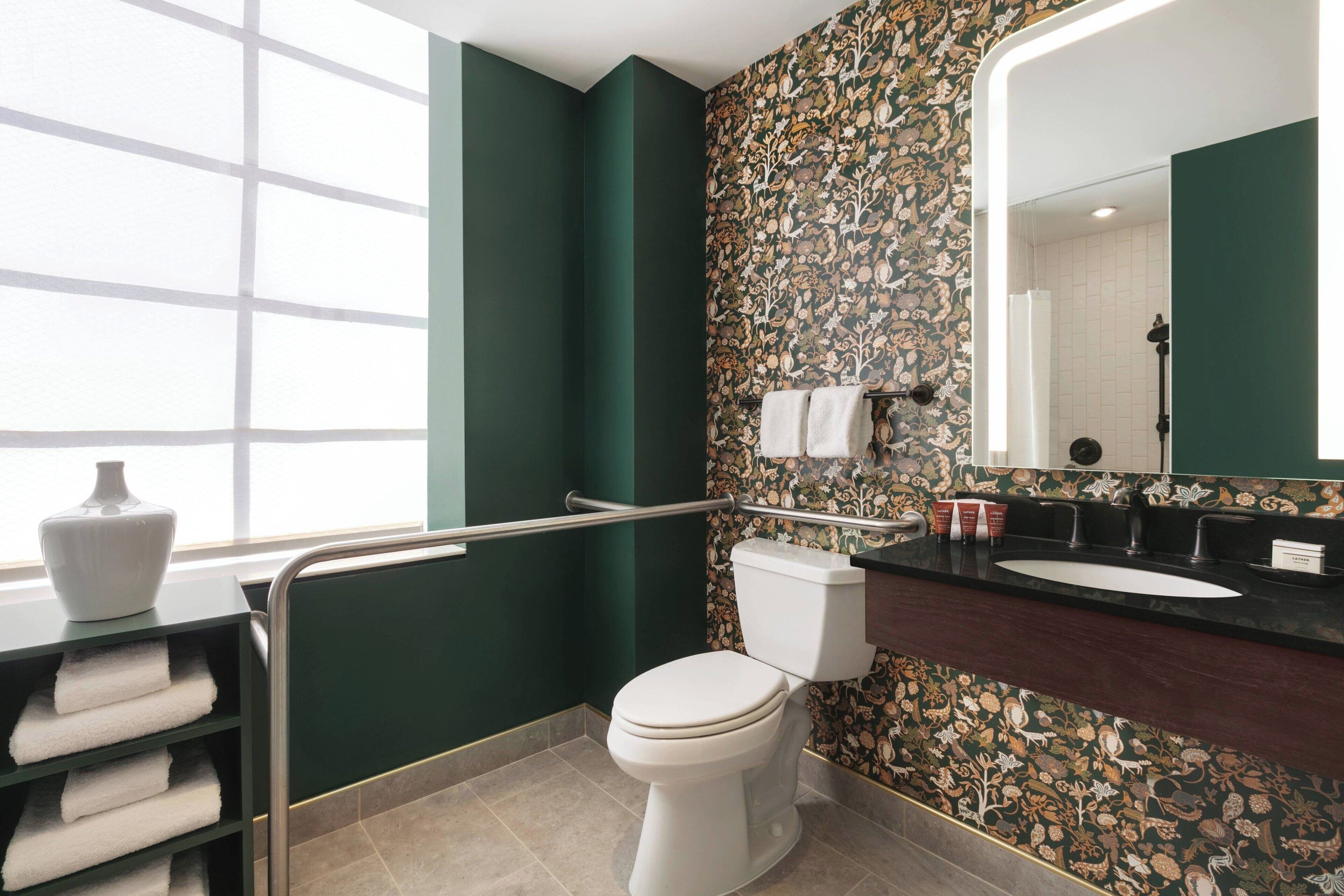 Un baño moderno con inodoro, lavabo y tocador. Cuenta con una pared verde con motivos florales, una ventana y un perchero con toallas dobladas.