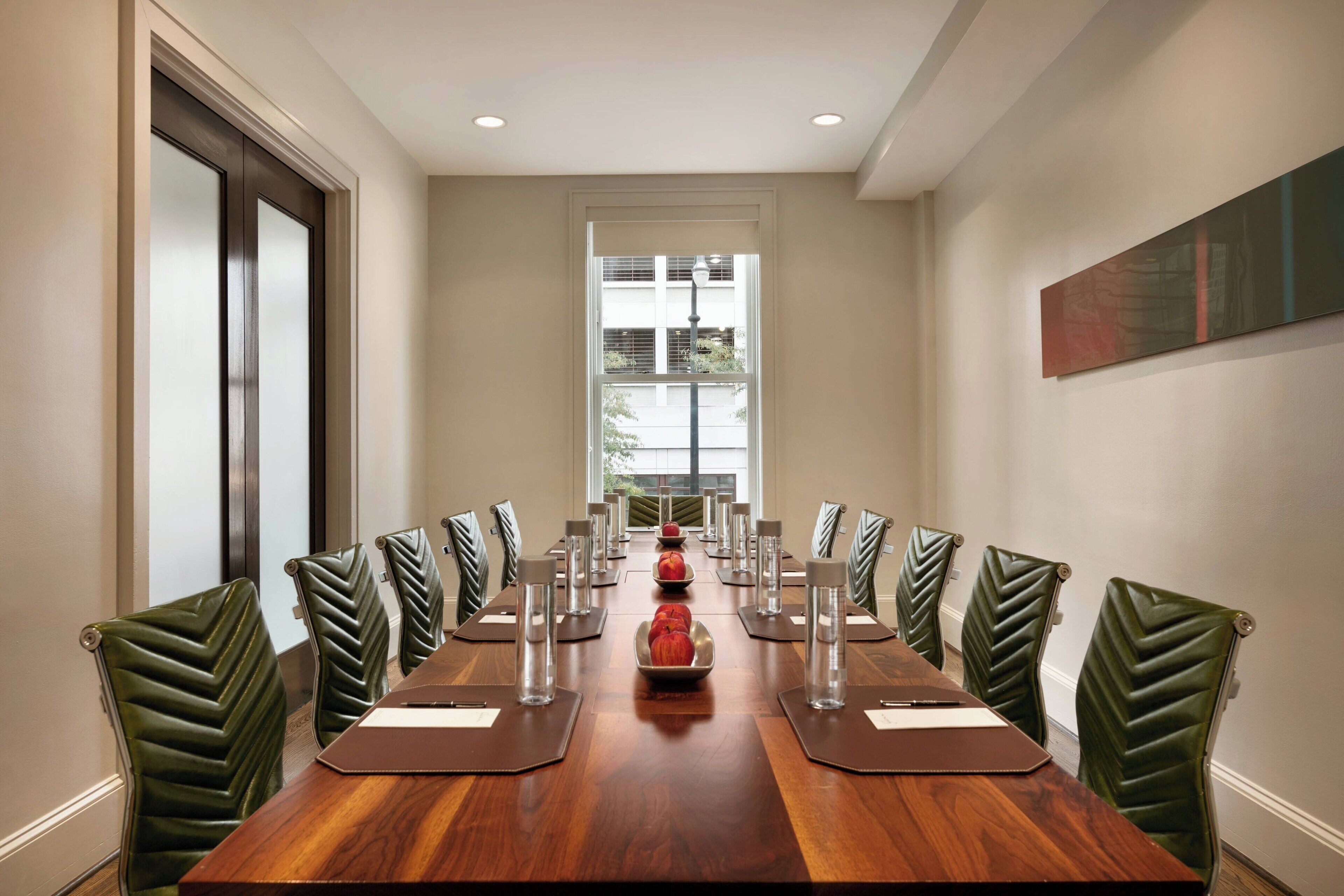 Een vergaderzaal met een lange tafel, groene bekleden stoelen en een groot raam met jaloezieën.