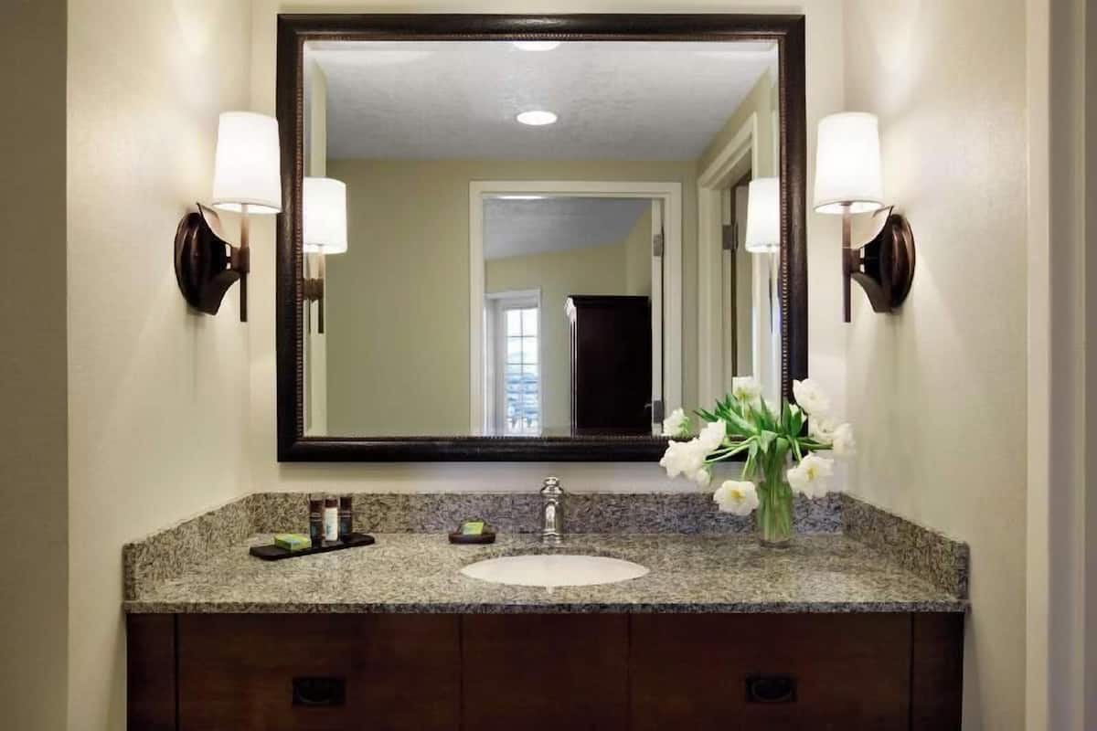 Une salle de bain dotée d’un comptoir en granit, d’un lavabo blanc, d’un grand miroir et de deux appliques murales.