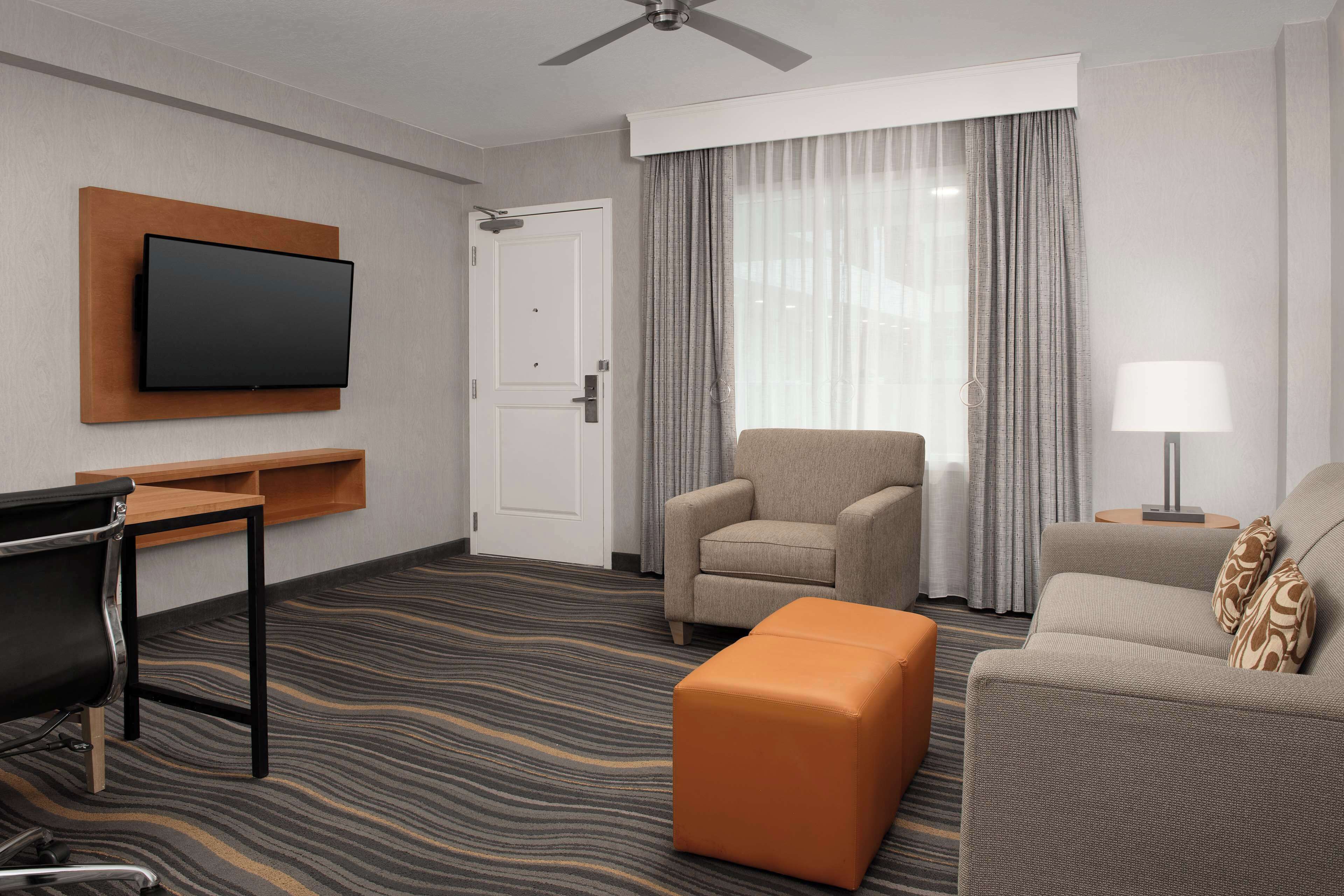 A hotel room with a flat-screen TV mounted on a wooden panel, a sofa, a chair, an ottoman, a desk with a chair, and a lamp.