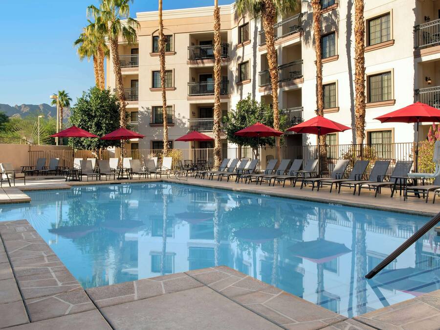 A swimming pool with lounge chairs and red umbrellas, surrounded by tall palm trees and a multi-story building with balconies.