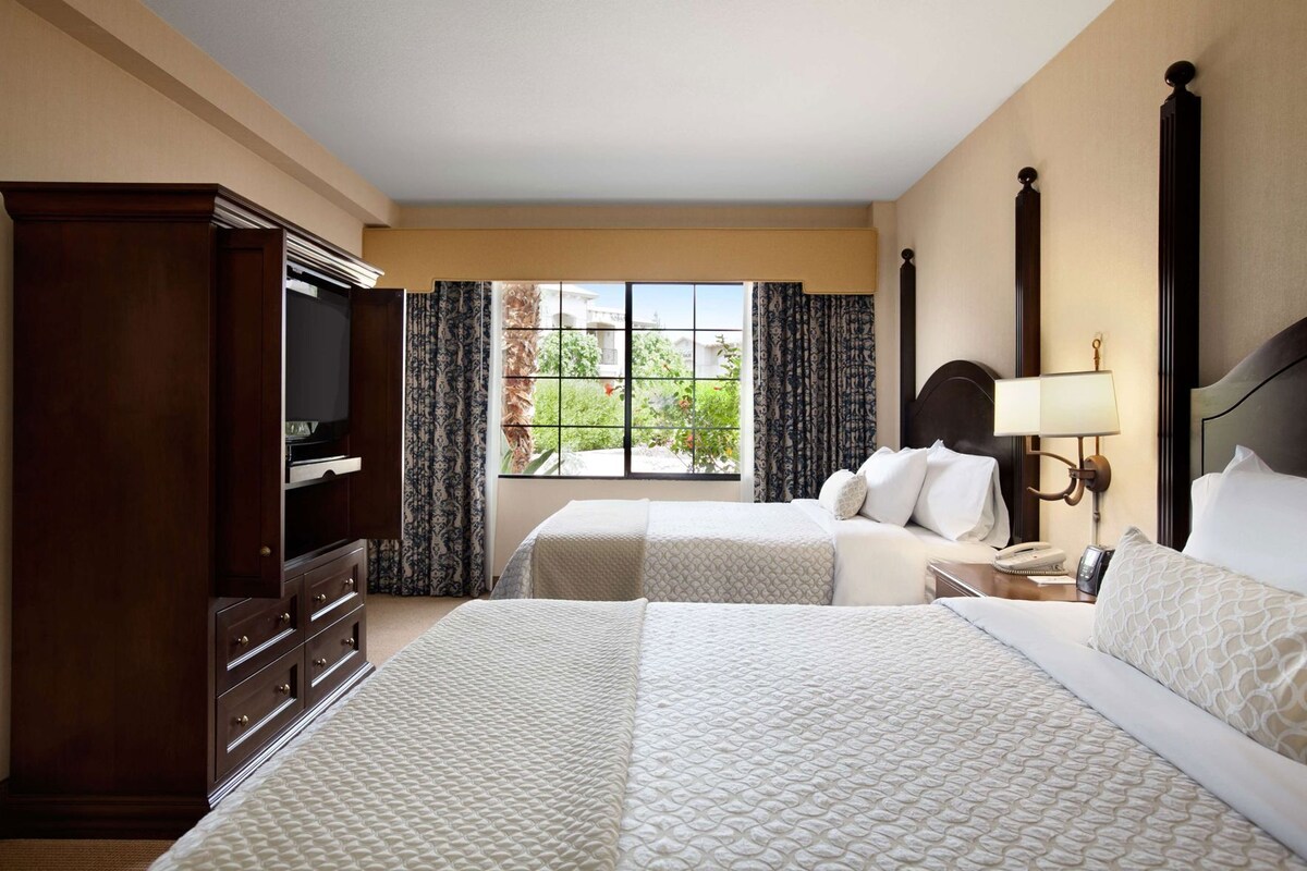 A hotel room with two beds, a wardrobe, a television, a window with curtains, and a nightstand with a lamp.