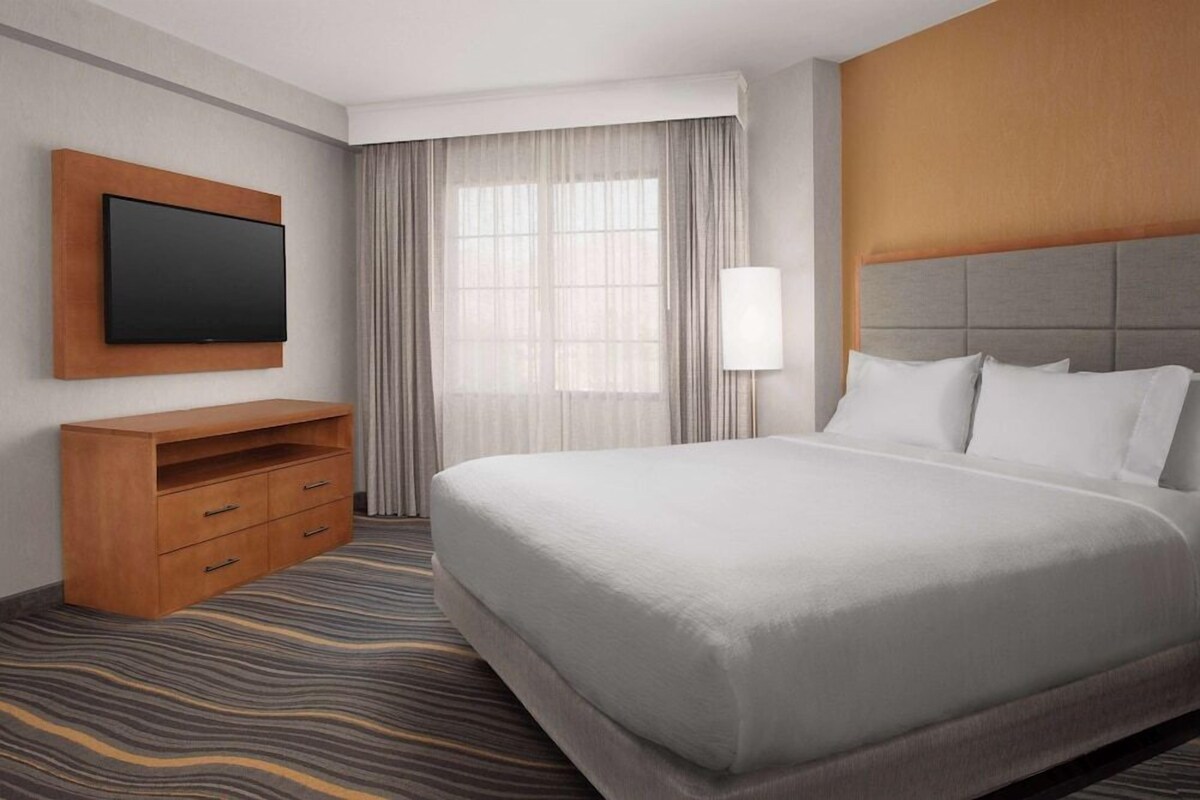 A hotel room with a large bed, a flat-screen TV mounted on a wooden panel, a bedside table with drawers, a lamp, and a window with sheer curtains.