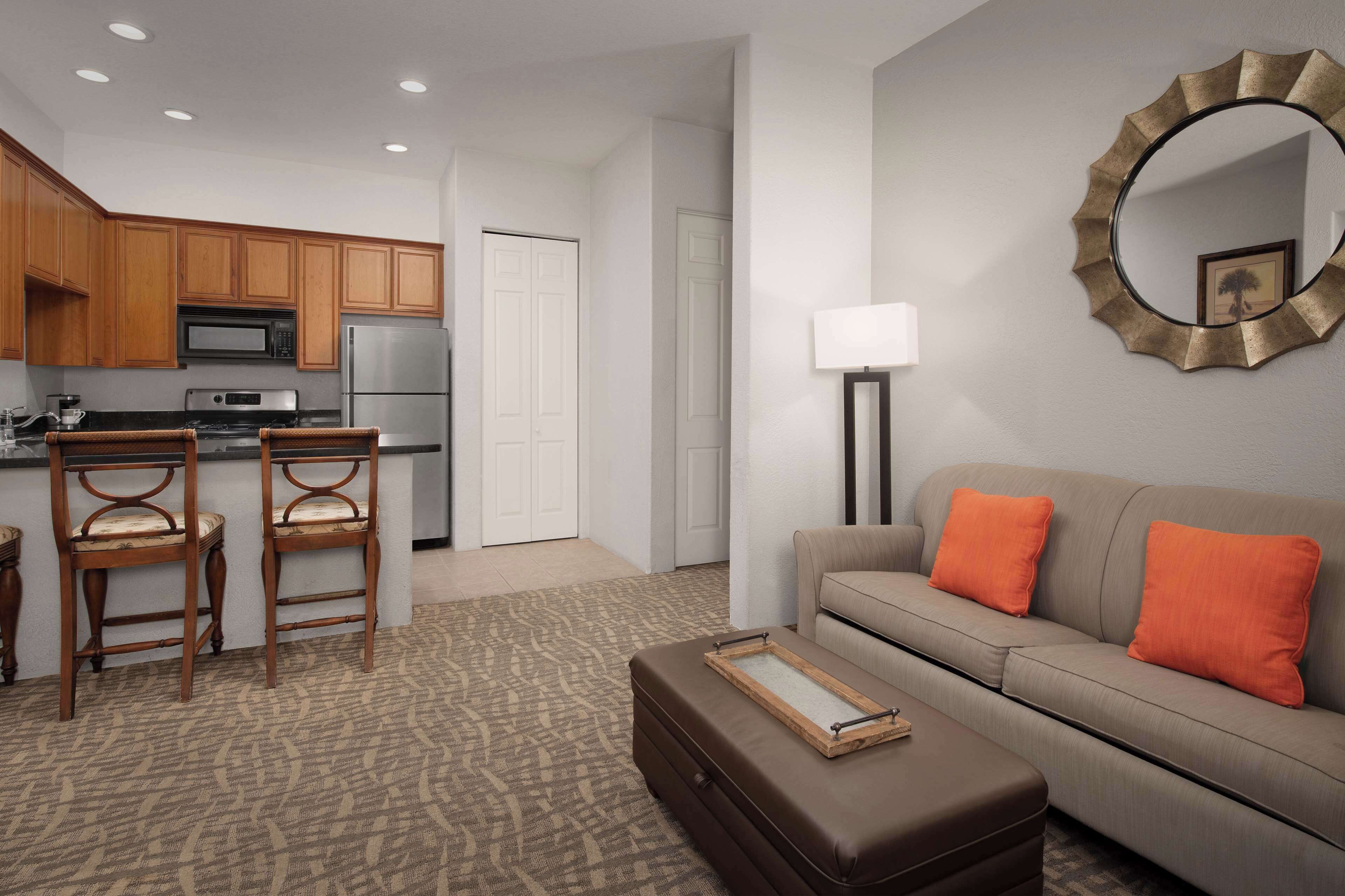 A modern living room with a sofa, ottoman, and chairs. There is a kitchen area with wooden cabinets and appliances.