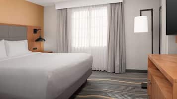 A hotel room with a large bed, a wooden dresser, a nightstand, a lamp, and a large window with curtains.