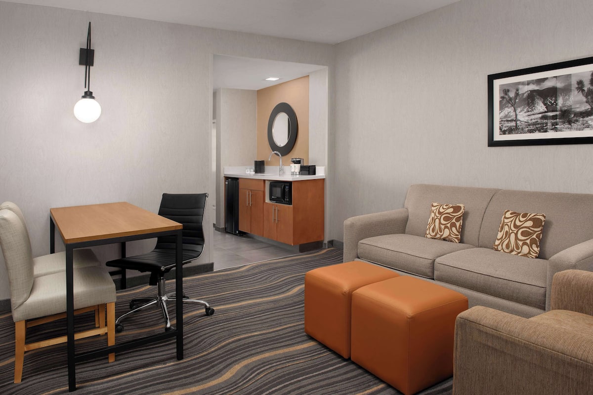 Une chambre d’hôtel équipée d ’une table à manger, d’un fauteuil de bureau, d’un canapé, d’un pouf, d’une kitchenette et d’un tableau encadré au mur.