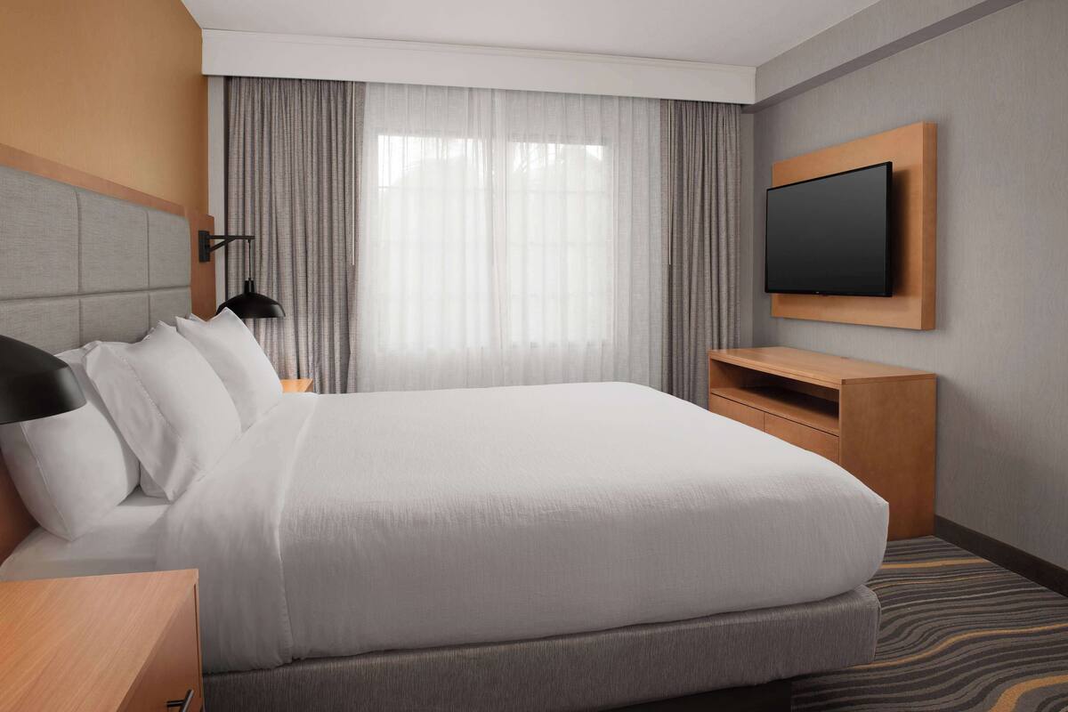 Une chambre d’hôtel avec un grand lit, un téléviseur fixé au mur, un bureau avec une lampe et une fenêtre avec des rideaux.