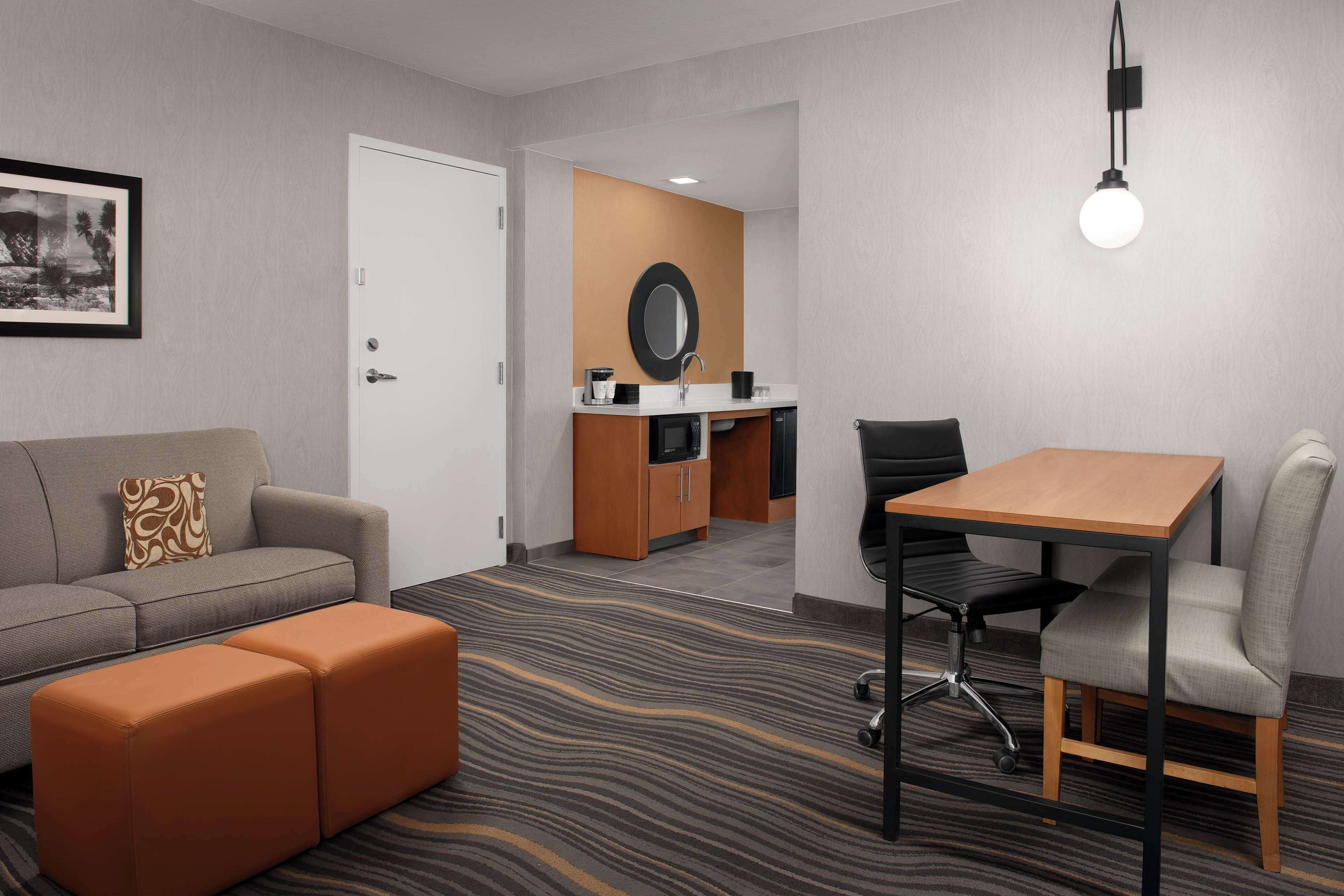 A hotel room with a sofa, ottoman, a small table with a chair, a desk with a chair, a mirror, and a light fixture.