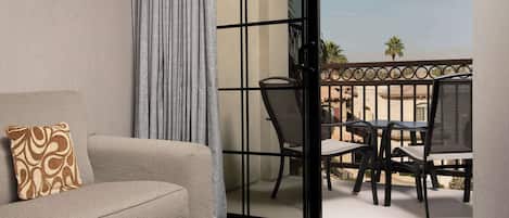 A hotel room with a sofa, an ottoman, a balcony with a table and chairs, and a view of palm trees.