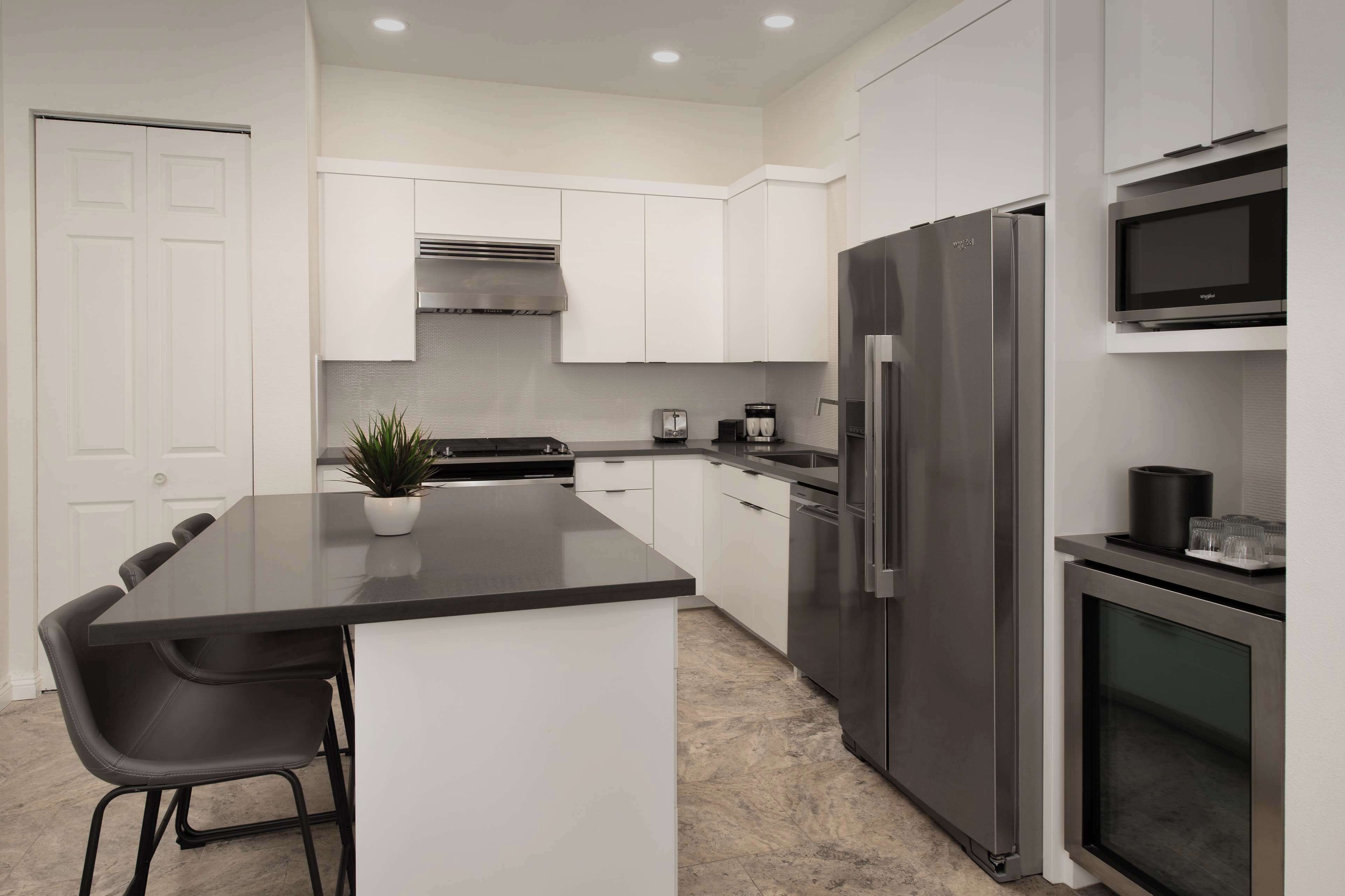 A modern kitchen with white cabinets, a stainless steel refrigerator, a built-in microwave, and a small island with bar stools.