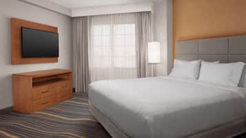 A hotel room with a large bed, a flat-screen TV mounted on a wooden panel, a bedside table with drawers, a lamp, and a window with sheer curtains.