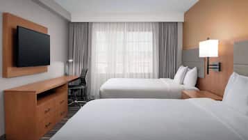 A hotel room with two beds, a TV mounted on a wooden panel, a desk with drawers, and a large window with sheer curtains.