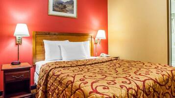 A hotel room with a wooden headboard, two bedside lamps, a bed with a patterned bedspread, a nightstand with a clock, and a framed picture on the wall.