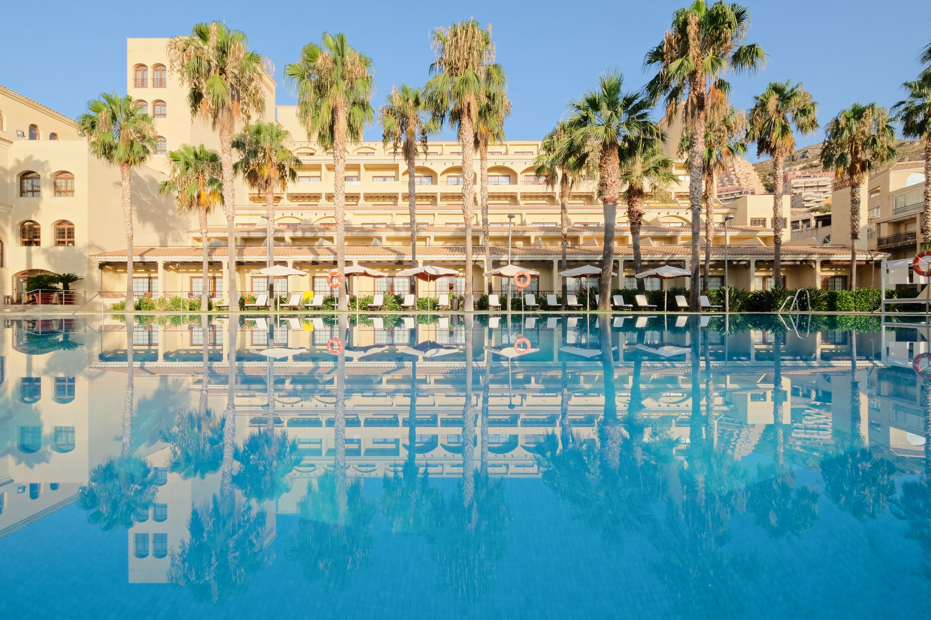 A hotel with a pool, palm trees, and a clear blue sky.