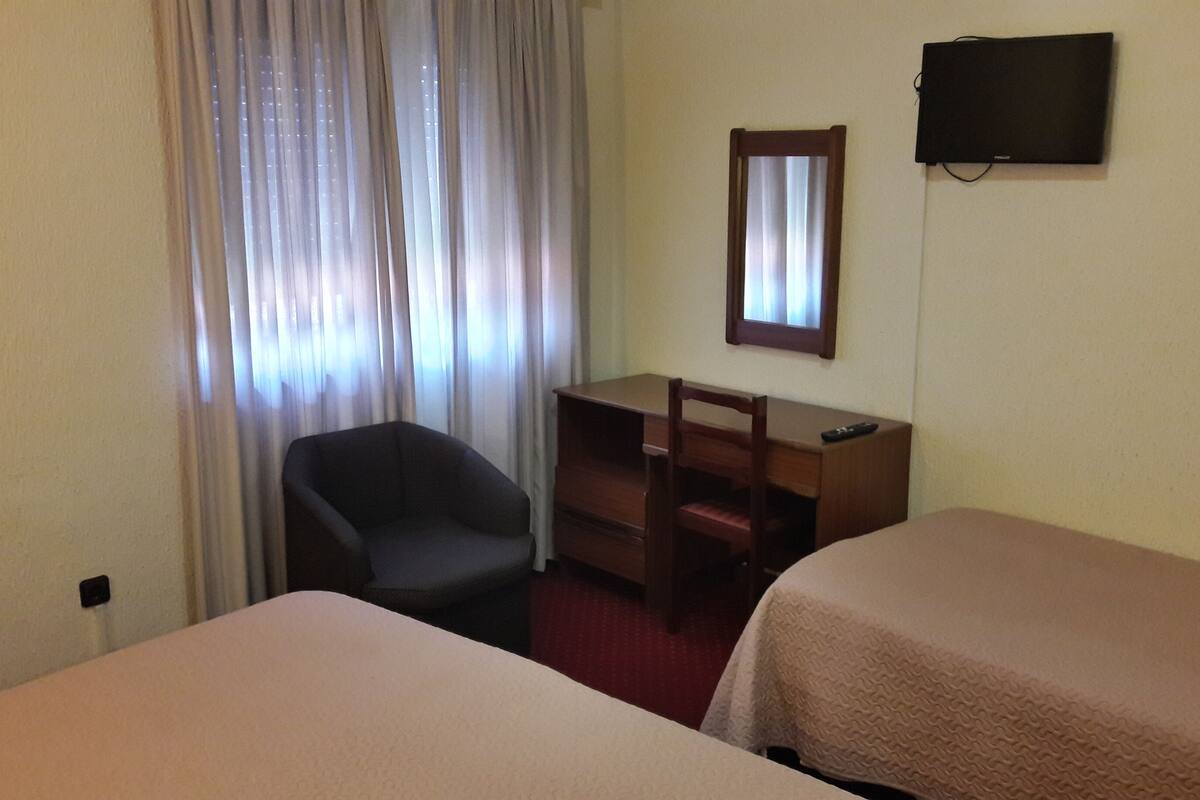 A hotel room with two single beds, a desk with a chair, a TV on the wall, and a window with curtains.