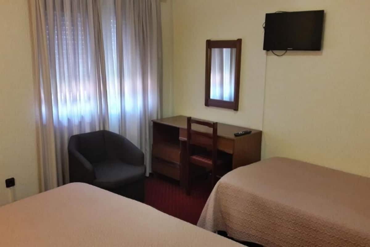 A hotel room with two beds, a desk, a chair, a TV, and a window with curtains.