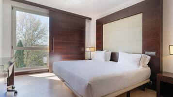 Una habitación de hotel moderna con una cama grande, un escritorio y vistas a árboles a través de un ventanal amplio.