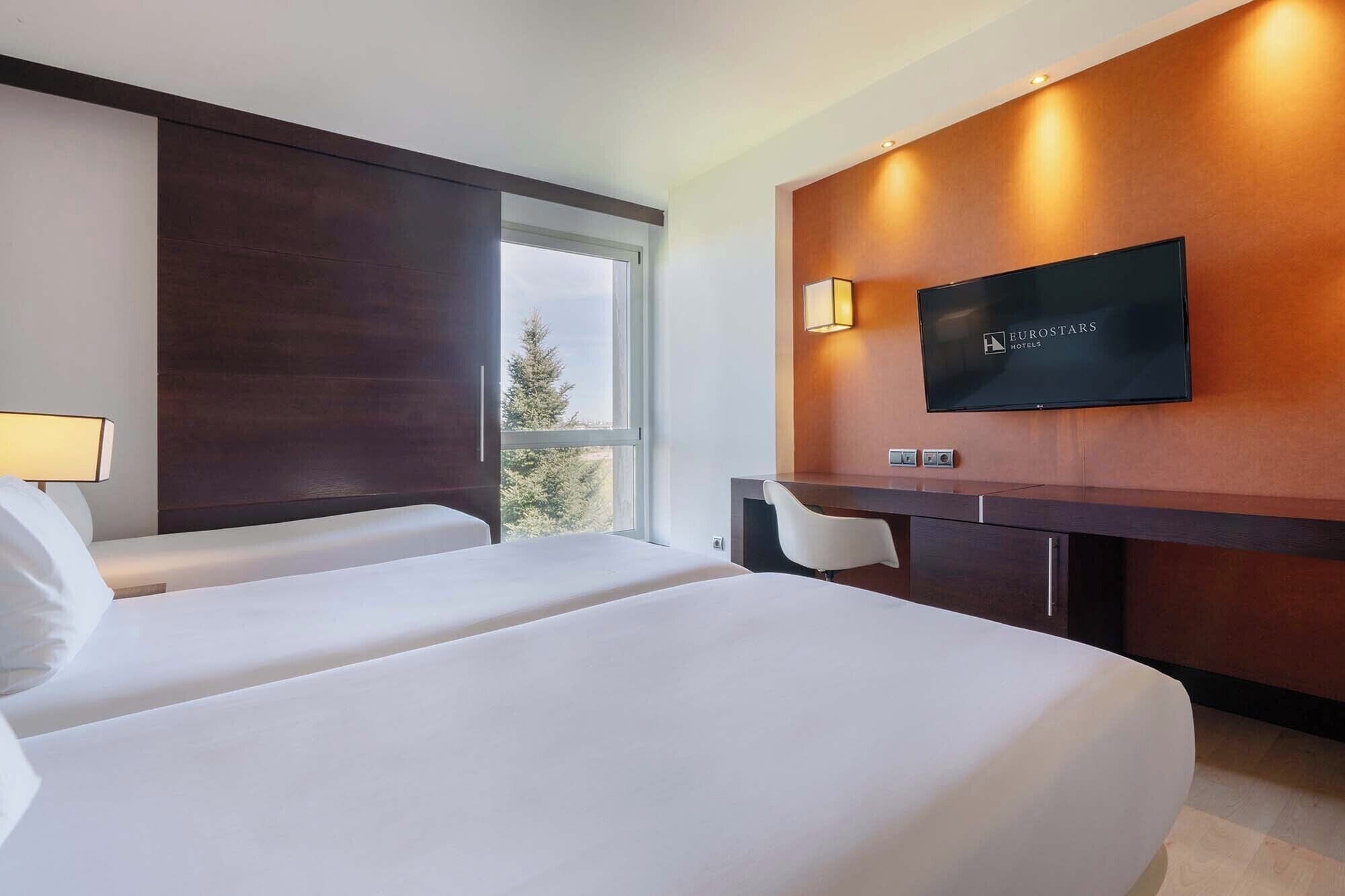 Quarto de hotel com duas camas, uma escrivaninha, televisão e janela com vista.