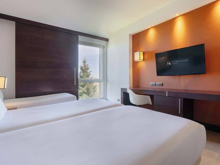 Quarto de hotel com duas camas, uma escrivaninha, televisão e janela com vista.