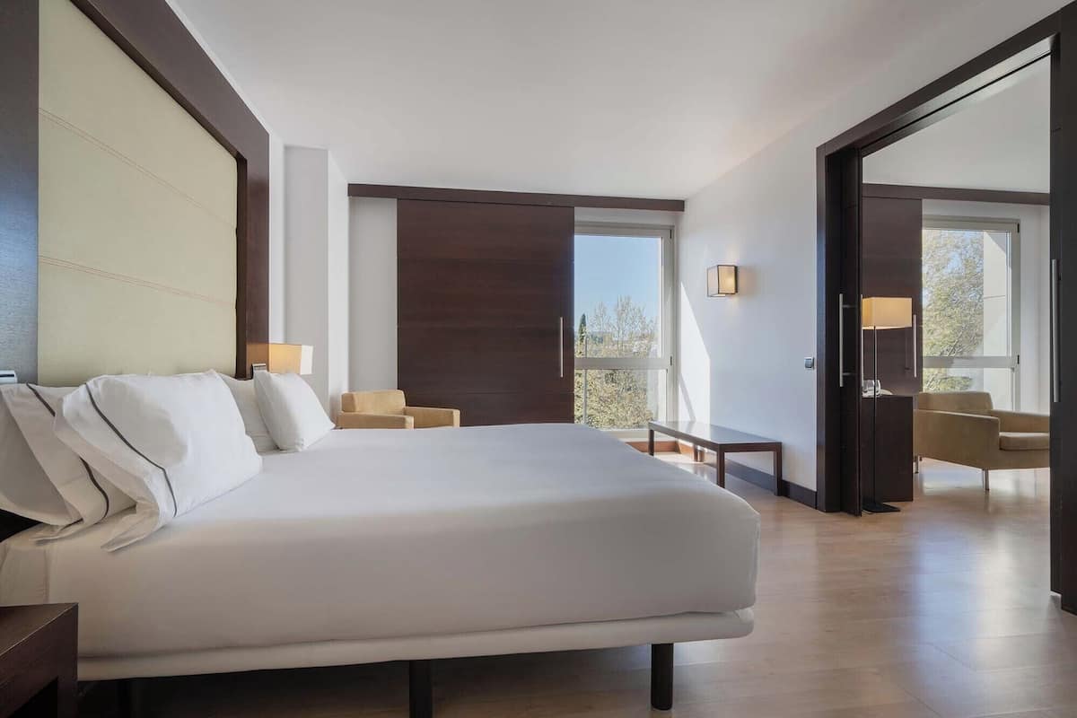 A hotel room with a large bed, a small table, two chairs, a balcony with a view, and a sliding door.