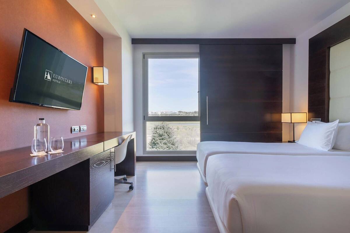 A hotel room with a bed, a desk with a TV, a window with a view, and a lamp.