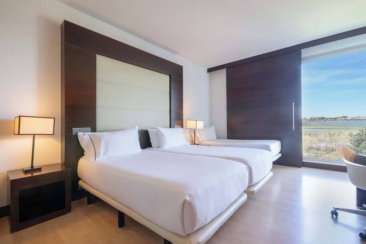 A hotel room with two beds, a desk, a chair, and a large window offering a view.