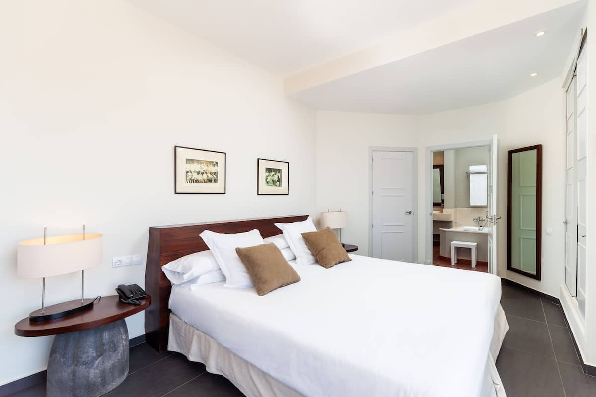 Ein Hotelzimmer mit einem großen Bett, Nachttischen mit Lampen, einem Telefon, gerahmten Bildern an der Wand und einem durch eine offene Tür einsehbaren Badezimmer.
