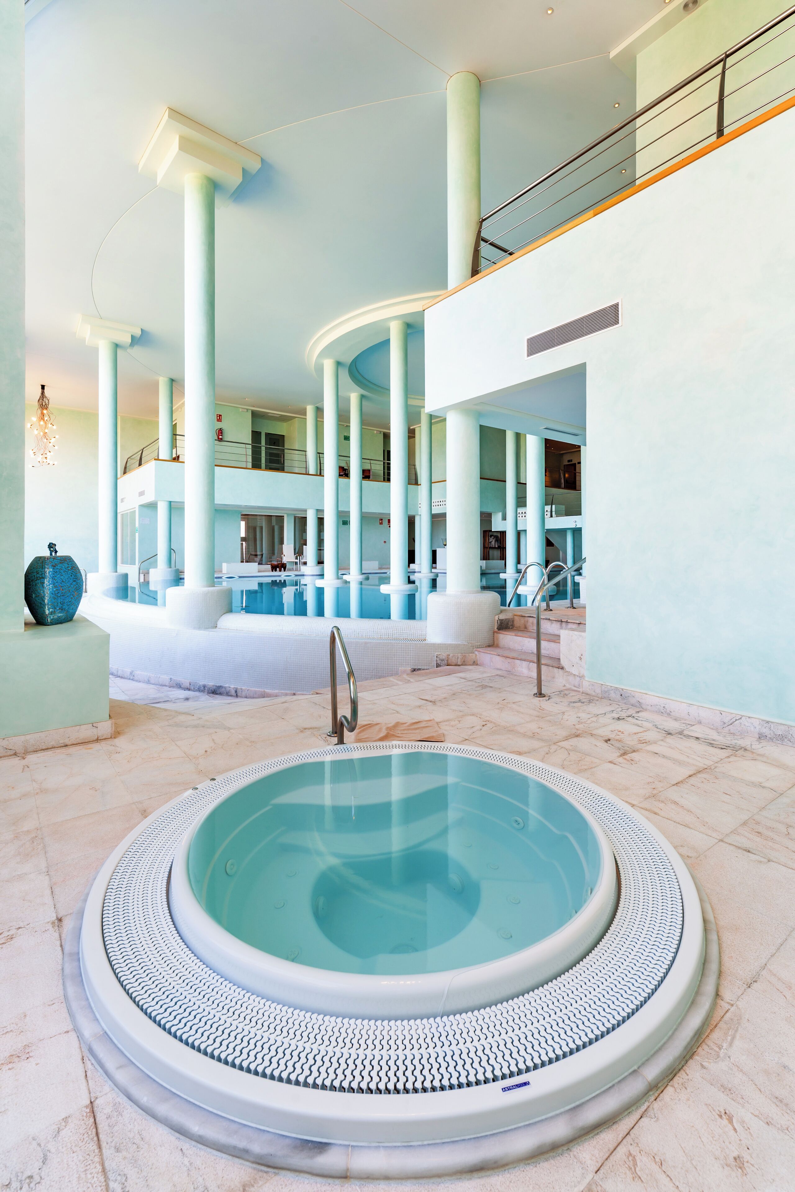 A spacious indoor swimming pool area with a central hot tub, surrounded by high ceilings and large columns.
