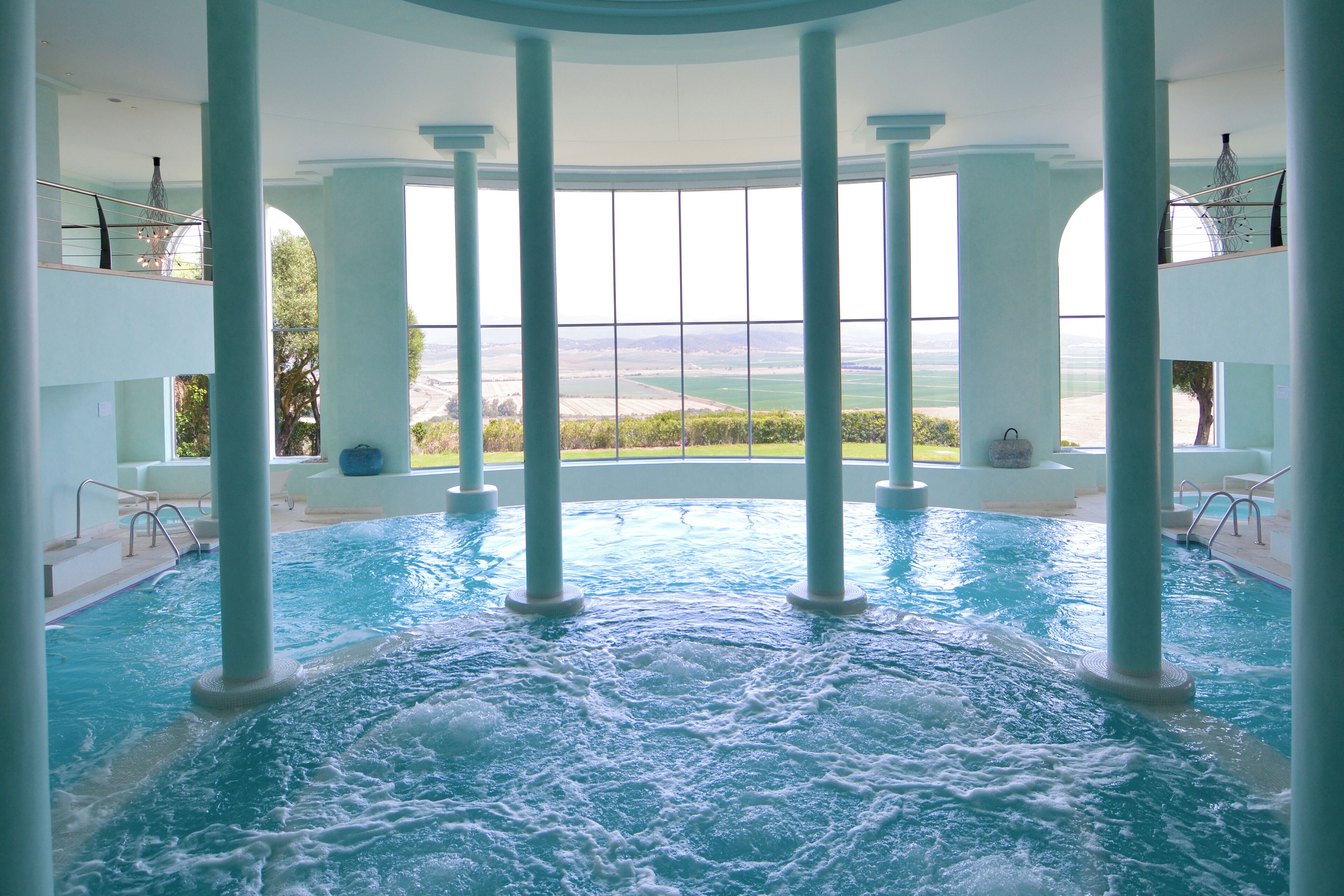 An indoor swimming pool with clear views of the outdoors, surrounded by columns and a high ceiling.