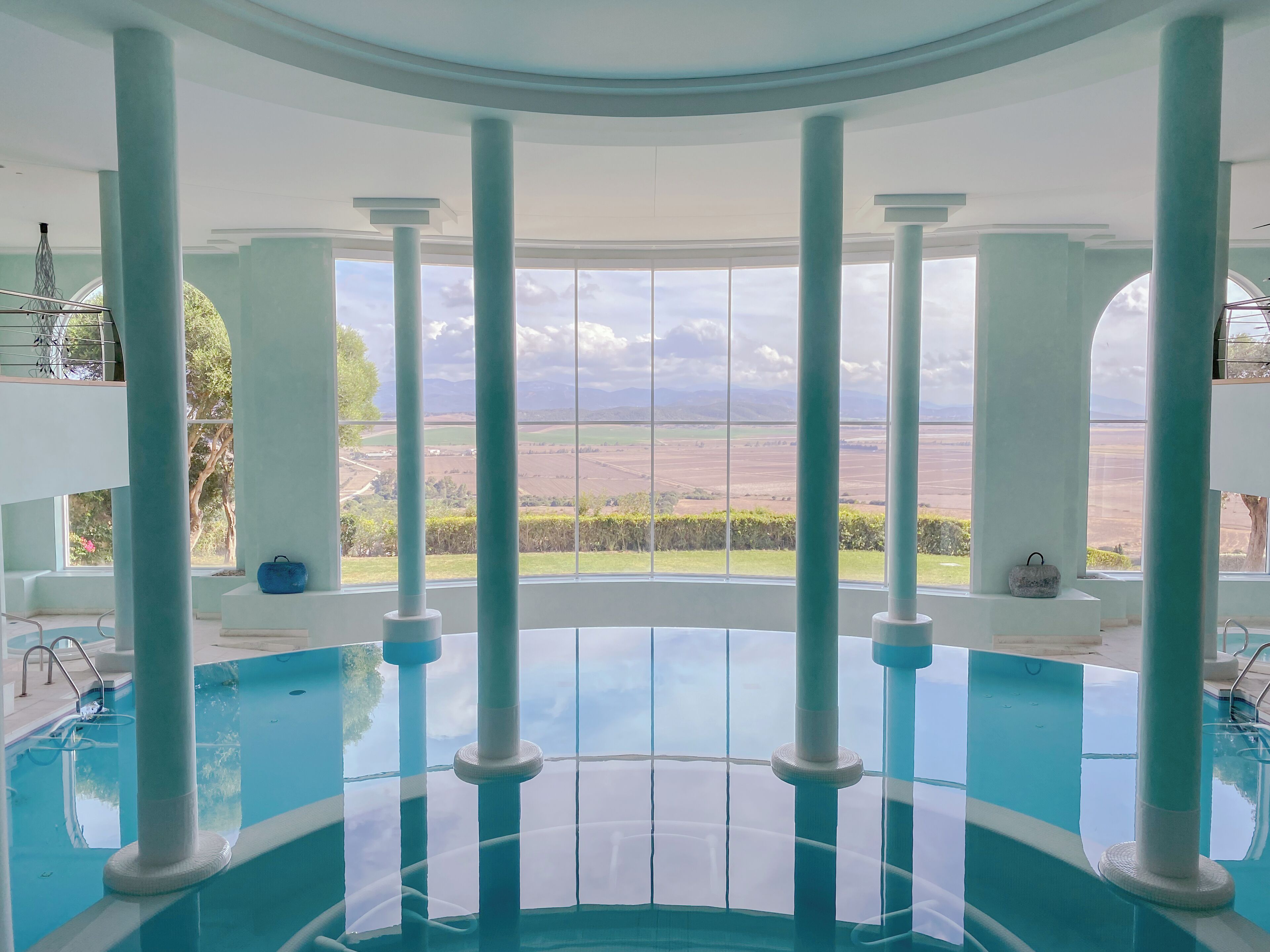 Zona de piscina con vistas a un paisaje a través de amplios ventanales, sostenida por columnas.
