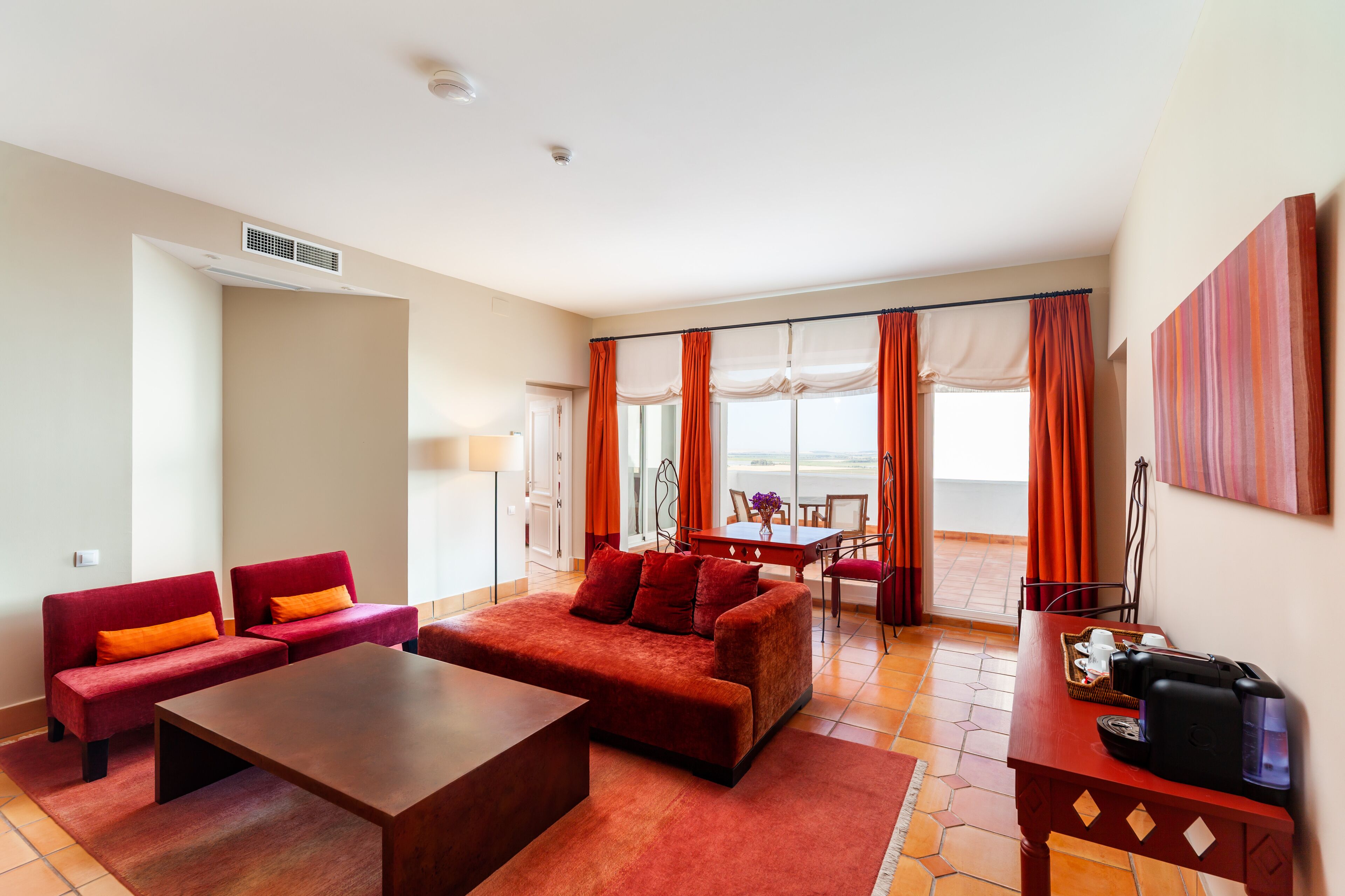 A hotel room with a red sofa set, a wooden coffee table, a dining area with a table and chairs, and a large window with curtains.