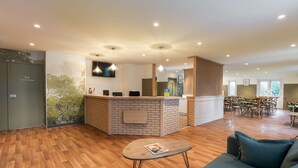 Un espace d’accueil moderne comprenant un comptoir de réception en bois, un coin salon avec un canapé et une petite table, ainsi qu’un espace repas avec une grande table et des chaises.