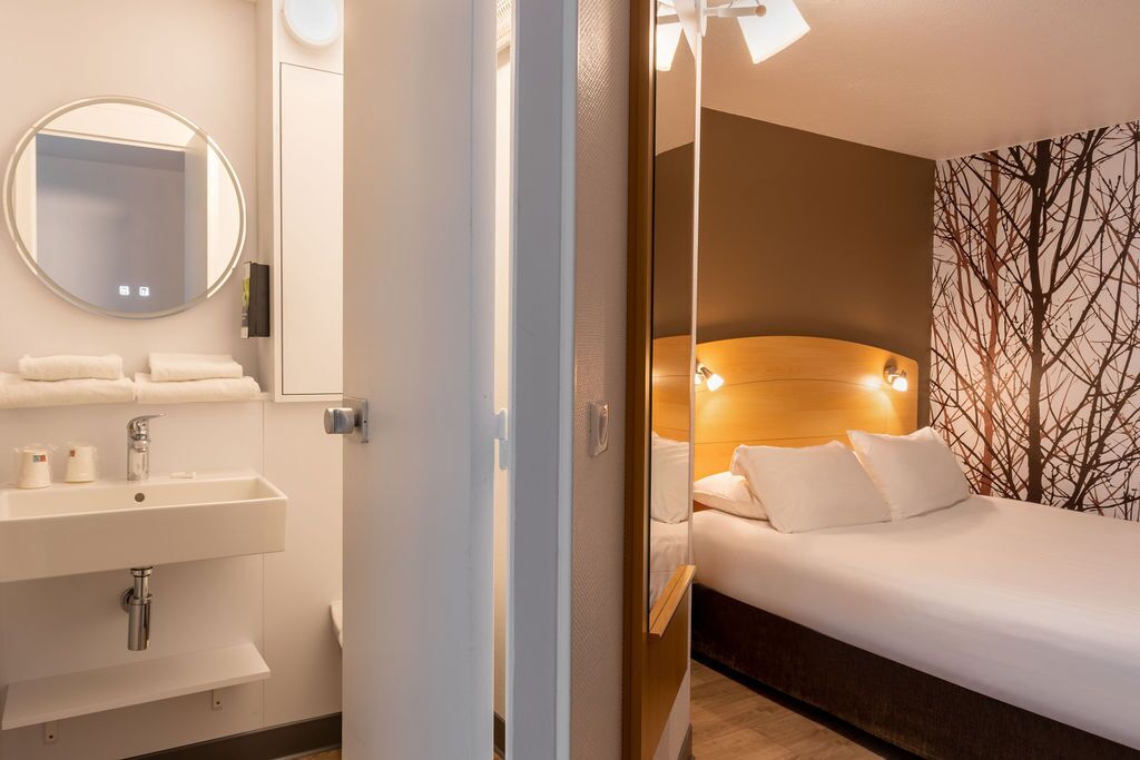 Habitación de hotel con cama, mesita de noche, espejo y baño con lavabo y toallero.