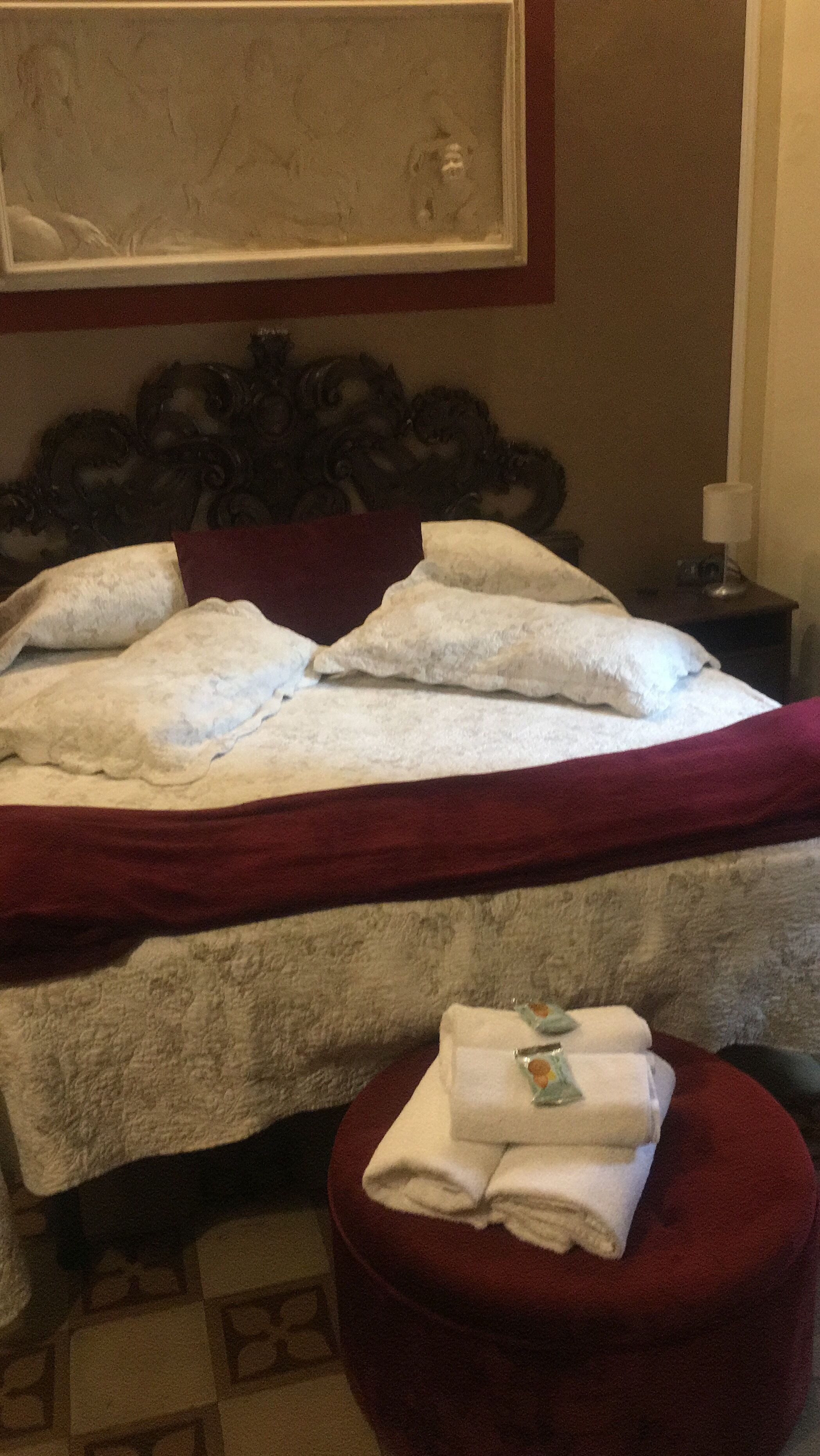 Una cama bien hecha con sábanas blancas y una colcha roja, un pequeño reposapiés redondo con toallas dobladas, y una obra de arte enmarcada sobre la cama.