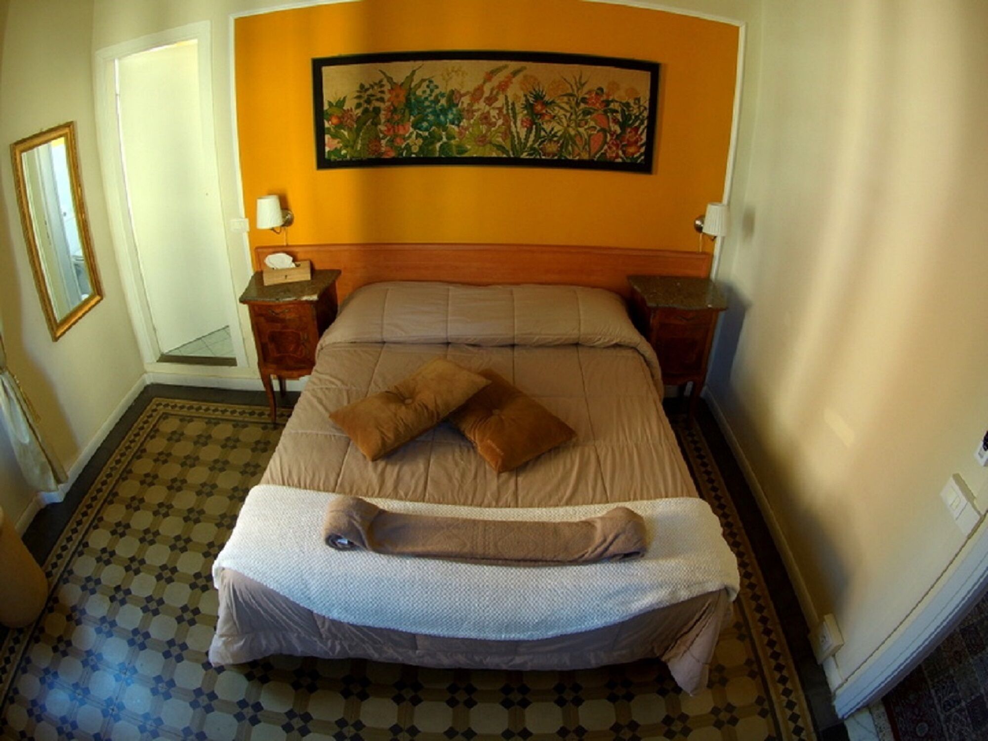 A hotel room with a bed, two bedside tables, a nightstand, a painting on the wall, and a patterned floor.