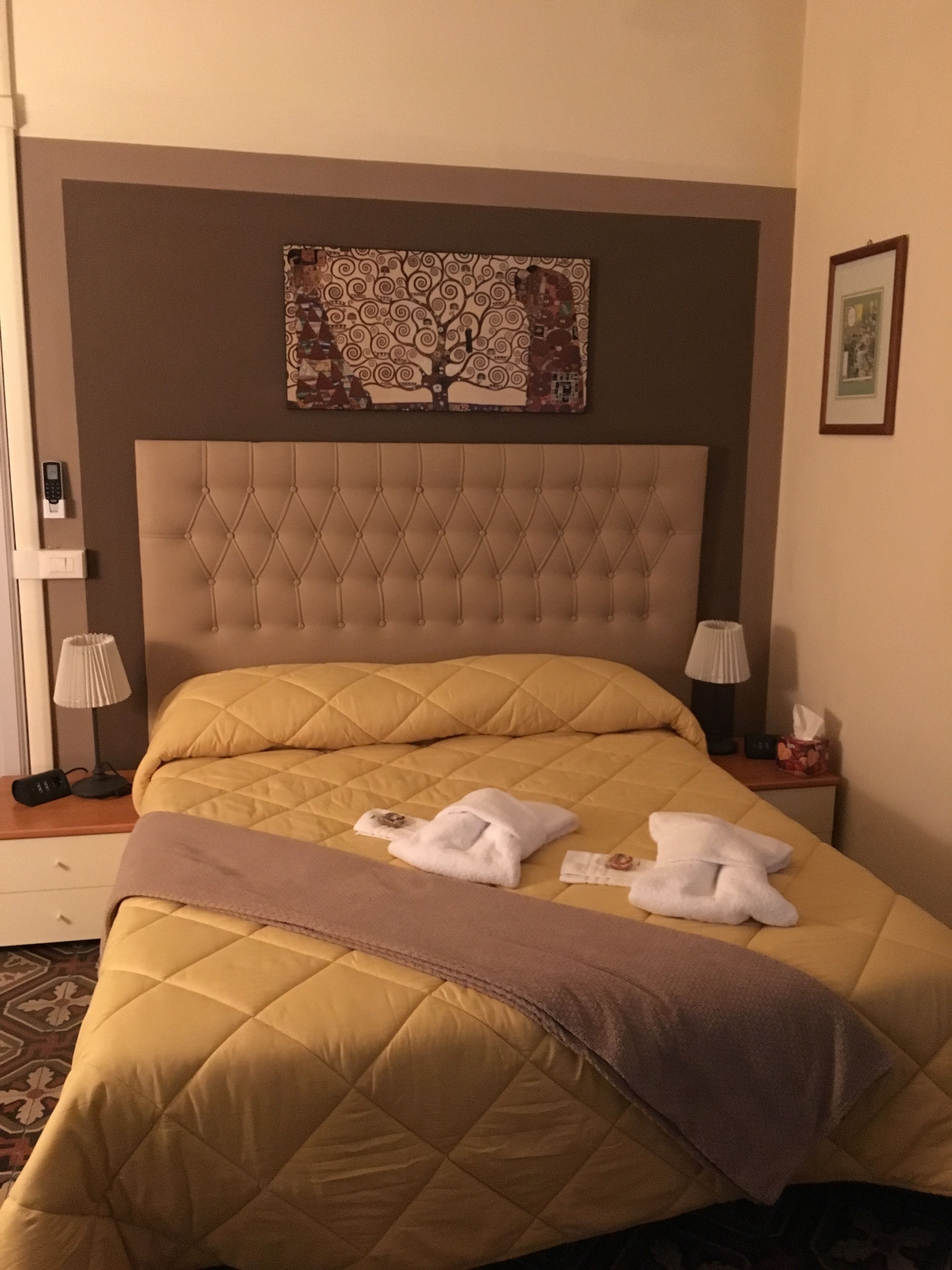 A hotel room with a large bed, a headboard with a decorative panel, two bedside lamps, and a bedside table with a lamp and a tissue box.