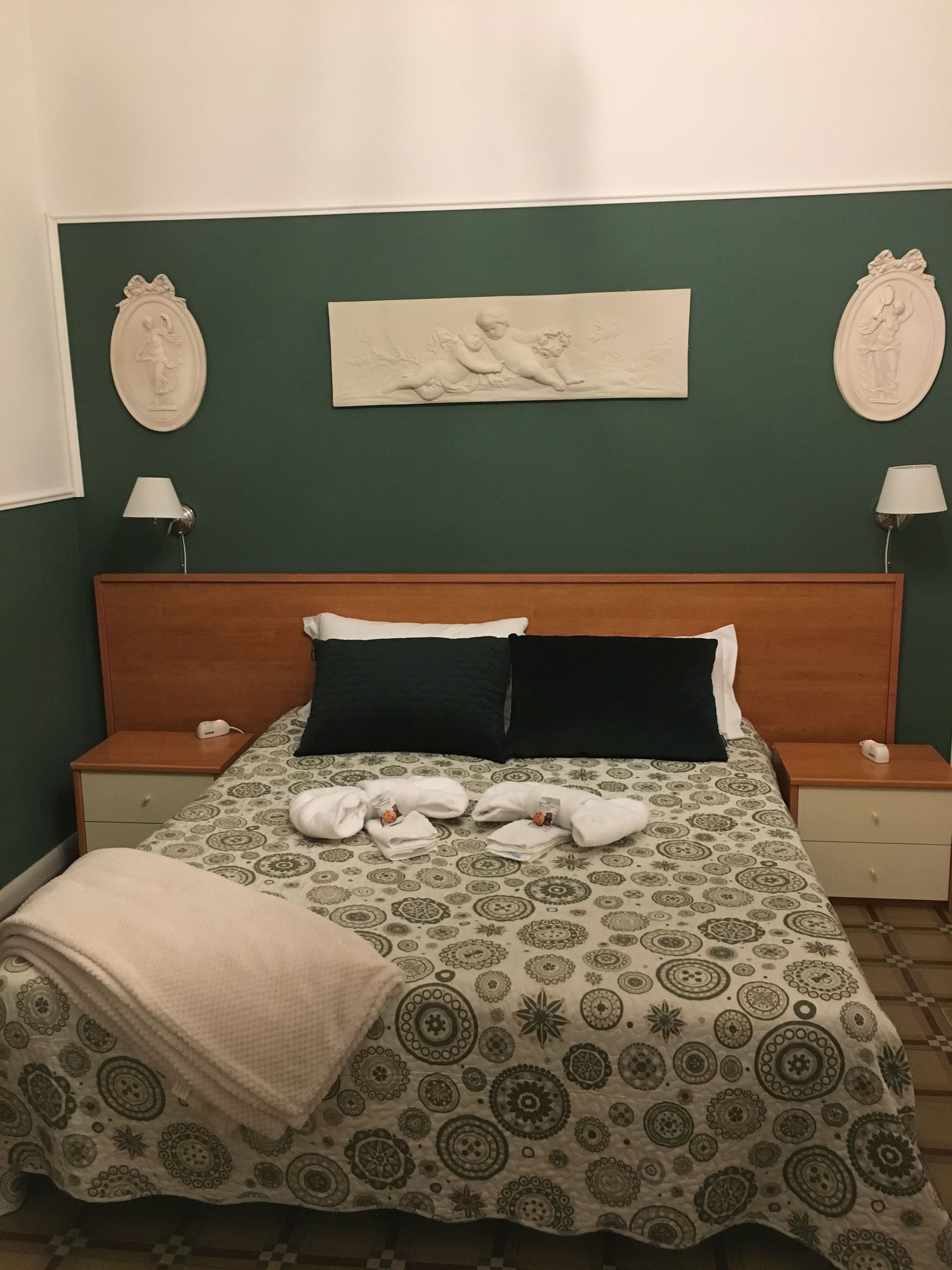Un lit bien fait, recouvert d’une couvre-lit à motifs, avec deux oreillers et une couverture. On y trouve deux tables de chevet avec des lampes, une œuvre d’art accrochée au mur et un mur peint en vert.