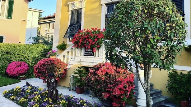 A yellow building with a flag, a staircase, and a garden with flowers and a tree. - Lucca in Villa San Donato (Lucca)
