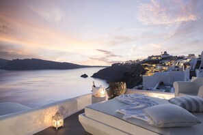 A sunset view from a balcony with white furniture, a stone wall, and a view of a coastal town. - Katikies Santorini – Gea House – The Leading Hotels of the World (Santorini)