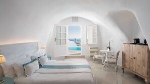 A bedroom with a bed, a desk, a chair, a cabinet, and a view of the sea. - Katikies Santorini – Gea House – The Leading Hotels of the World (Santorini)