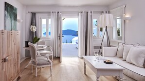 A modern, well-lit room with a white sofa, a dining table, and a view of the sea. - Katikies Santorini – Gea House – The Leading Hotels of the World (Santorini)
