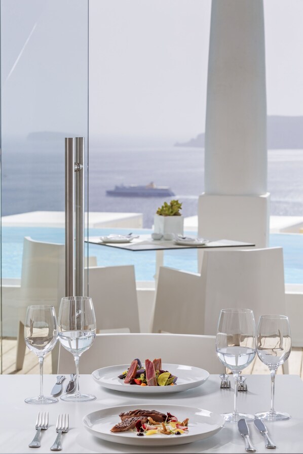 A dining table with two plates of food, two wine glasses, and cutlery. A tall, slender column is visible in the foreground. The background features a view of the sea and a ship.
