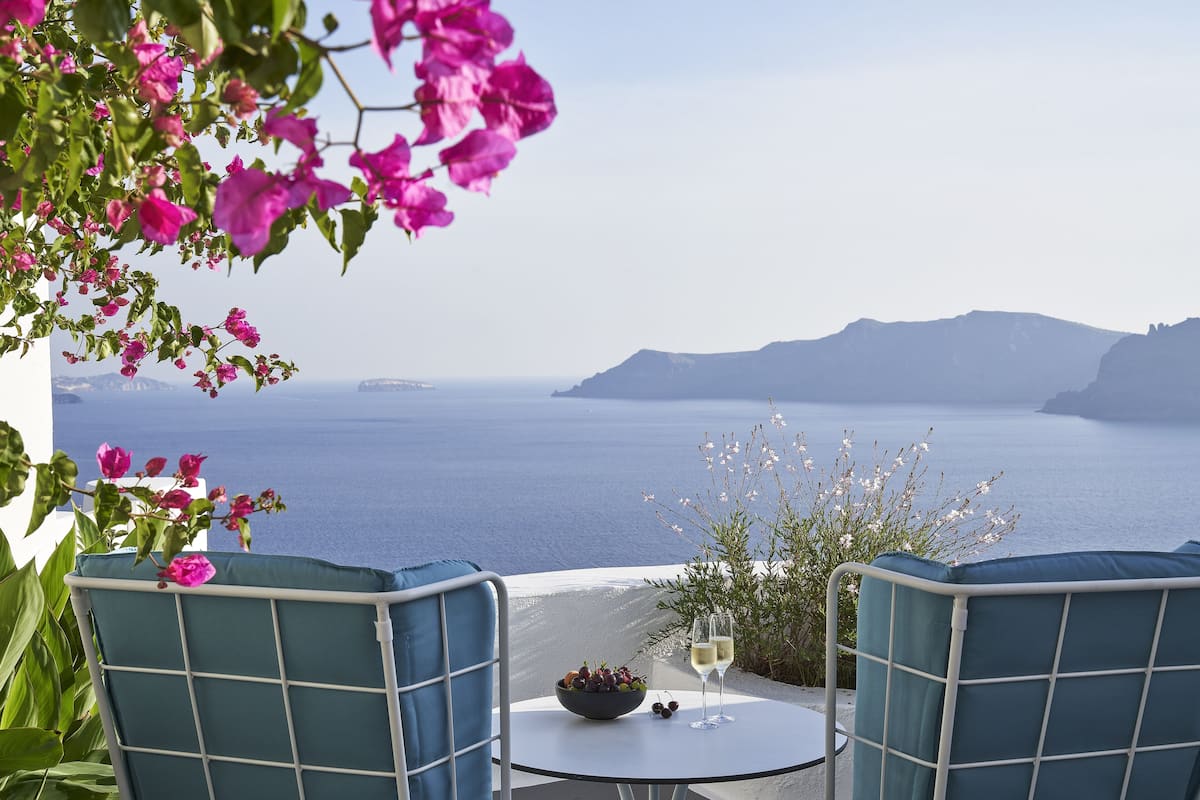 Una mesa con un bol de fruta y dos copas de vino, con vista al mar y montañas al fondo.
