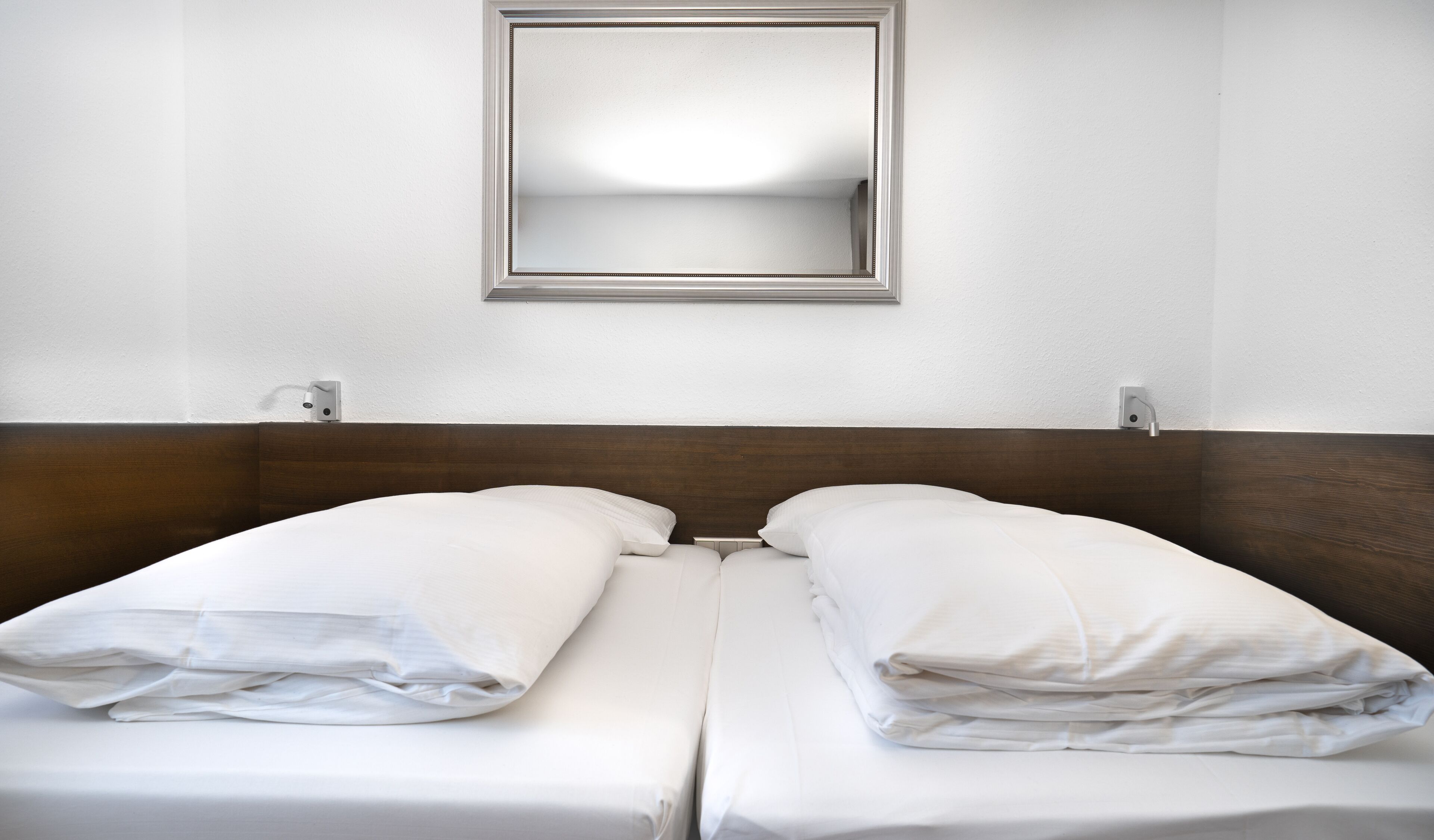 A hotel room with a wooden headboard, two white beds with pillows, a mirror on the wall, and a plain white wall.