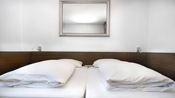 Quarto de hotel com cabeceira de madeira, duas camas brancas com almofadas, um espelho na parede e uma parede branca lisa.